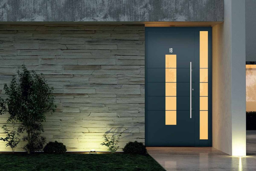 A charcoal aluminium entrance door in the evening with ambient garden lighting and a bush